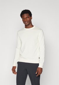 Maglione lavorato a maglia color crema chiaro con collo rotondo, maniche lunghe e polsini a costine. Presenta un piccolo etichetta con logo sul lato inferiore. Indossato con pantaloni scuri.