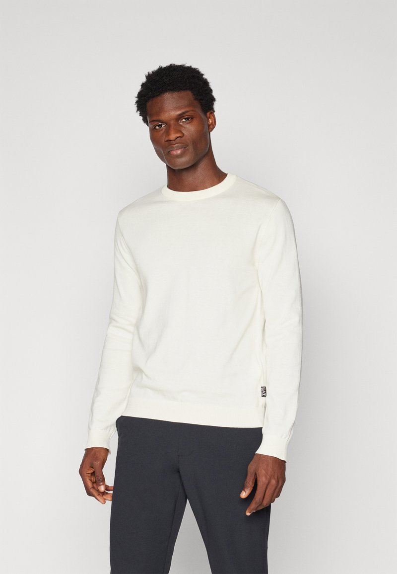 Maglione lavorato a maglia color crema chiaro con collo rotondo, maniche lunghe e polsini a costine. Presenta un piccolo etichetta con logo sul lato inferiore. Indossato con pantaloni scuri.