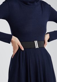 Svart elastiskt bälte med silverdetaljer, med horisontella ränder, bärs över en marinblå klänning med hög hals och långa ärmar.