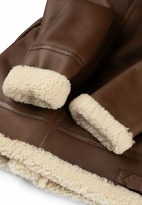 Braune Lederjacke mit Lammfellmanschetten und -futter, glatter Textur und stabilen Nähten. Auffälliger Kontrast zwischen braunen und cremefarbenen Tönen.