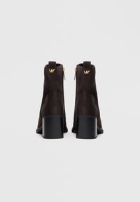 Stivaletti alla caviglia in suede marrone scuro con tacco quadrato, dettagli in oro e zip posteriori. Texture liscia e design pulito e minimal.