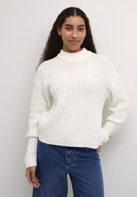 Maglione bianco a coste con colletto alto, maniche lunghe e vestibilità morbida, abbinato a pantaloni in denim blu.