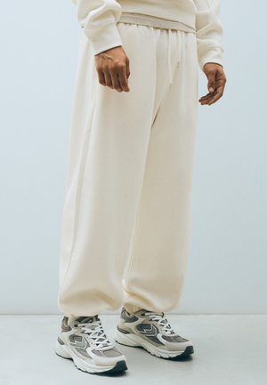 Personne portant un pantalon de survêtement crème ample et des baskets athlétiques gris et blanc, debout sur un sol clair.