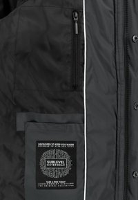 Black puffer jacket featuring a zippered chest pocket, white piping, and a fabric label on the lower pocket with textured lettering.