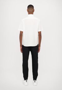 Chemise blanche à manches courtes avec un ourlet droit, associée à un pantalon noir fuselé et des baskets blanches avec des accents noirs au niveau du talon.