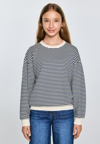 Dryžuota džemperis tamsiai mėlynos ir kreminės spalvų, ilgomis rankovėmis ir su gumuotais rankogaliais. Suderintas su mėlynomis džinsinėmis kelnėmis, pasižyminčiomis laisvu siluetu ir tiesiomis kojomis.