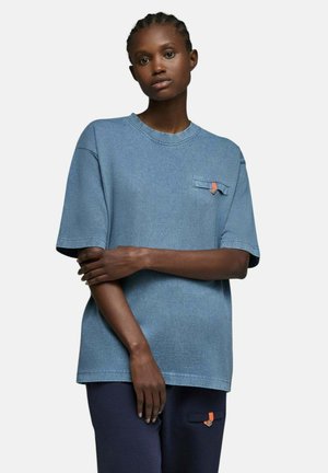 Femme aux cheveux tressés, portant un t-shirt bleu ample et un pantalon sombre, debout les bras croisés contre un fond clair uni.