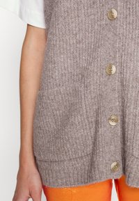Gros plan d'une personne portant un cardigan beige en tricot sans manches avec de gros boutons sur une chemise blanche et un pantalon orange vif.