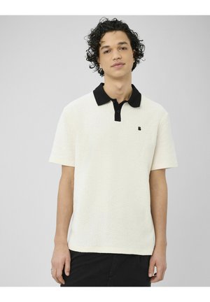 Jeune homme aux cheveux bouclés portant un polo blanc à manches courtes avec un col noir et un pantalon noir, debout devant un fond gris.