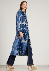 Abrigo largo azul con un patrón floral texturizado, combinado con pantalones de mezclilla oscuros acampanados y tacones beiges, sobre un fondo neutro.