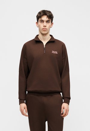 Młody mężczyzna w brązowej bluzie z krótkim zamkiem i dopasowanych spodniach z logo "the new originals" na piersi i udzie.