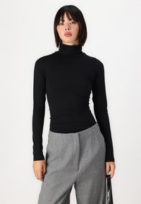 Crna dugah rukava turtleneck majica, uskog kroja, izrađena od glatkog materijala, uparena sa visokim sivim nabranim hlačama. Minimalistički stil.