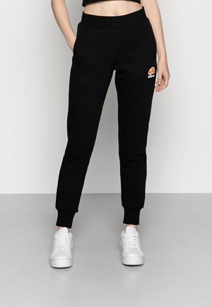 Schwarze Jogginghose mit geripptem Bund, elastischen Bündchen, Seitentaschen und einem mehrfarbigen gestickten Logo am linken Oberschenkel. Getragen mit weißen Sneakers.
