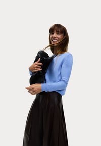 Ljusblå tröja med långa ärmar, kombinerad med en mörk plisserad kjol. Modellen håller en svart väska och ler mot en neutral bakgrund.
