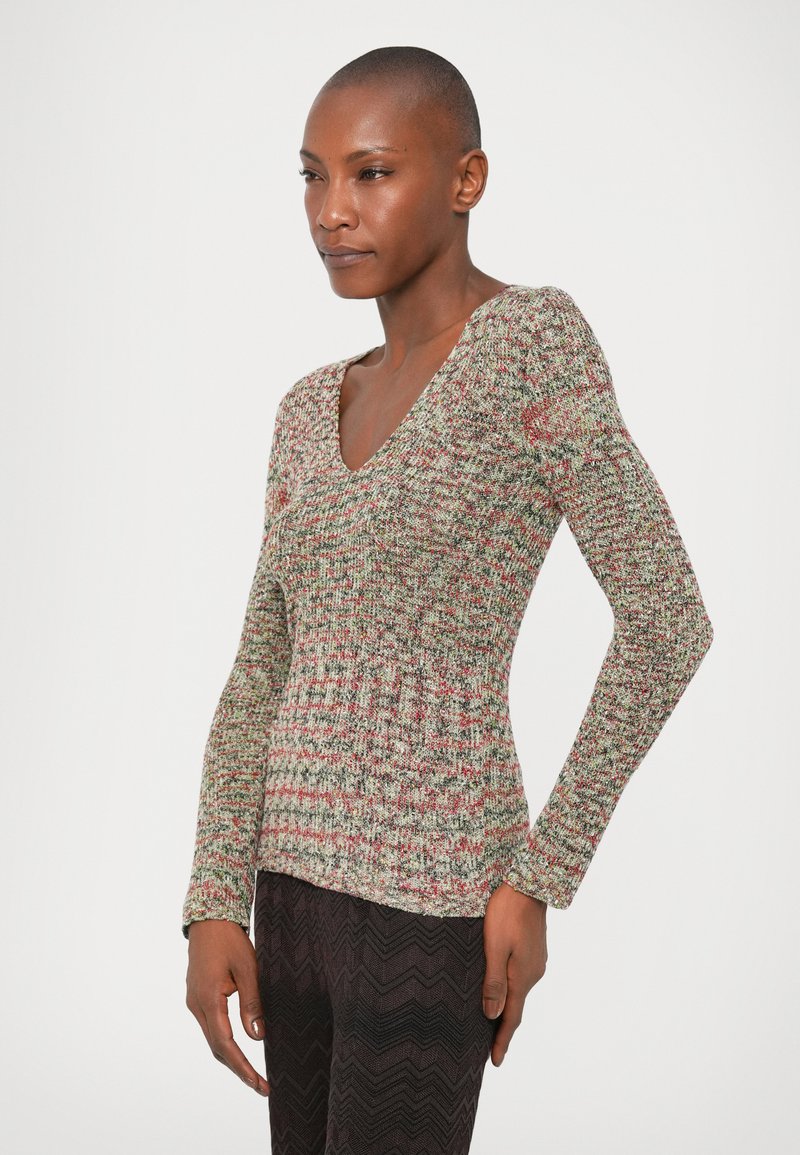 Langärmliger Strickpullover mit mehrfarbigem Muster, ausgestattet mit einem tiefen V-Ausschnitt und einer taillierten Passform. Getragen mit strukturierten, dunkel gemusterten Hosen.