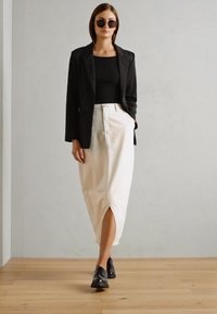 Blazer nero su una maglia nera, abbinato a una gonna midi color crema con uno spacco frontale e scarpe nere con lacci. Texture liscia, design minimale.
