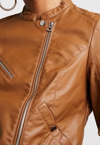 Hnědá kožená bunda s vysokým límcem, diagonalním zipem, patentním knoflíkem a šikmými kapsami. Hladká textura s jemným prošitím.