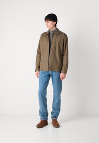 Olivengrønn knappet jakke, lys grå zip-up skjorte under, blå denimjeans, brune støvler og svart strikket lue. Enkle bakgrunner.