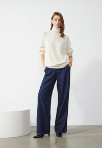 Pull en crêpe à col roulé et côtes associé à un pantalon large en denim bleu foncé et des bottines noires. Fond minimaliste.