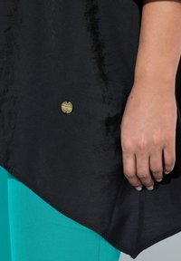 Top nero testurizzato con un'etichetta circolare dorata, indossato sopra pantaloni turchesi, con un orlo asimmetrico e una finitura di tessuto liscia.