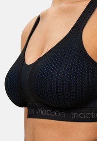 Brassière de sport noire avec un motif texturé composé de points bleus et une large bande noire portant le texte "traction". Les bretelles réglables offrent un maintien optimal.
