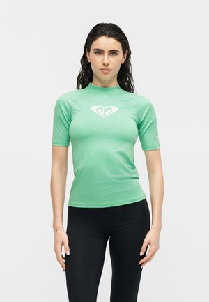 Žena s mokrom tamnom kosom koja nosi svijetlozeleni rash guard s kratkim rukavima, bijelim logotipom i crne tajice stoji na bijeloj pozadini.