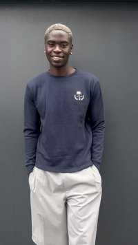 Giovane uomo con capelli biondi corti, che indossa un maglione blu navy con logo di palma e pantaloni chiari, sorridente con le mani in tasca.