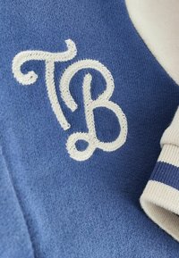 Blau gestrickter Stoff mit einem weißen gestickten Monogramm "TB" und dekorativem Steppmuster, neben einem strukturierten, beige gestreiften Bündchen.
