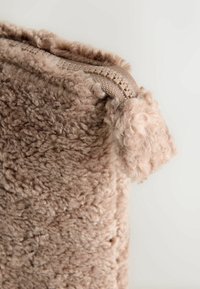 Beige fuzzy taske med en blød tekstureret overflade og en matchende lynlås med en plys trækflap mod en ensfarvet baggrund.