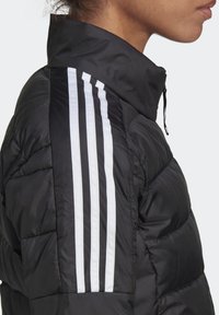 Černá prošívaná bunda s vysokým límcem a třemi bílými pruhy na rameni, vyrobená z hladkého, lesklého materiálu.