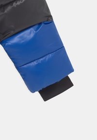 Rękaw pikowanej kurtki z czarnymi i niebieskimi sekcjami, z błyszczącą teksturą i elastycznym czarnym mankietem. Widoczne detale szwów na materiale.