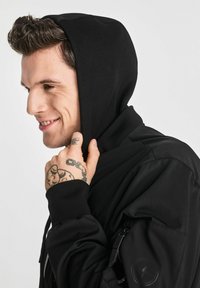 Czarna bluza z kapturem, z sznurkiem ściągającym i prążkowanymi mankietami, z zamykaną na zamek kieszenią na rękawie i naszywką z logo na ramieniu. Widoczny detal faktury materiału.