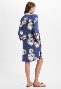 Blaues Leinenkleid mit weißem Blumenmuster, Dreiviertelärmel, seitlichen Taschen und Schlitzen. Flache Sandalen runden den Look ab.