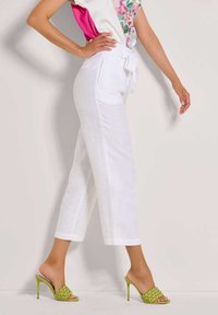 Witte linnen broek met een knoop in de taille, gecombineerd met groene gevlochten sandaals met hak. De outfit heeft een bloemenpatroon met roze accenten.
