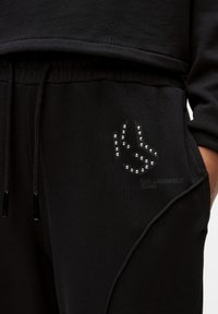 Pantaloni de trening negri dintr-un material moale, cu un elastic ajustabil la talie, buzunare laterale și un detaliu cu ținte pe coapsa stângă.