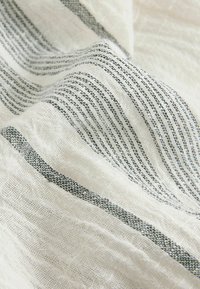 Weißer Stoff mit dünnen schwarzen Streifen, der eine strukturierte, gewebte Optik aufweist. Die Zusammensetzung scheint leicht und atmungsaktiv zu sein.