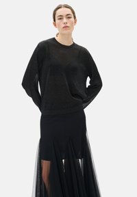 Femme portant un haut noir transparent à manches longues et une jupe noire avec des panneaux transparents, se tenant les mains derrière le dos.