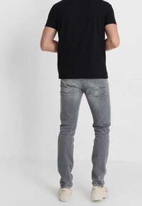 Svart kortärmad t-shirt, grå slim-fit jeans med bakfickor och subtila sömdetaljer, bärs med beige skor, sedd bakifrån.