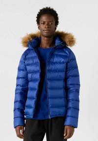 Joven vistiendo una chaqueta acolchada azul con capucha de piel, camisa azul debajo, de pie contra un fondo claro.