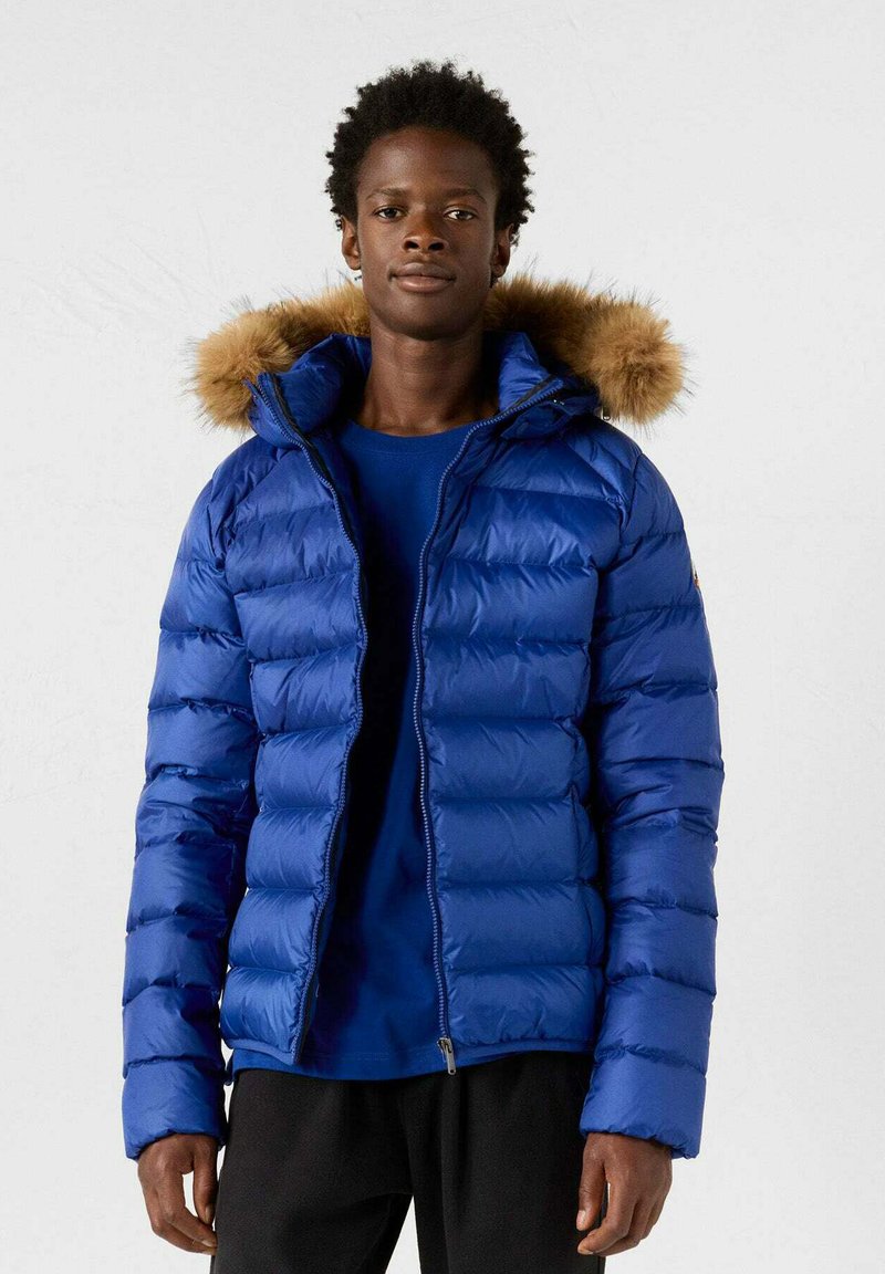 Jongeman in een blauwe pufferjack met een capuchon met bontafwerking, een blauw shirt eronder, staand tegen een effen lichte achtergrond.