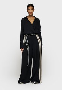 Zwarte langwerpige shirt met knoopdetail in de taille, gecombineerd met wijde zwarte pantalons met crèmekleurige verticale strepen en een logo-riem.