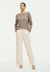 Pulover bej tricotat cu guler ribat, asortat cu pantaloni largi crem cu buzunare laterale și nasture. Modelul stă încrezător.