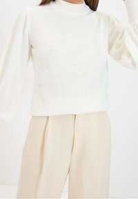 Pull blanc texturé avec un col montant, doté de manches volumineuses. Associé à un pantalon beige taille haute. Tissu en maille douce.