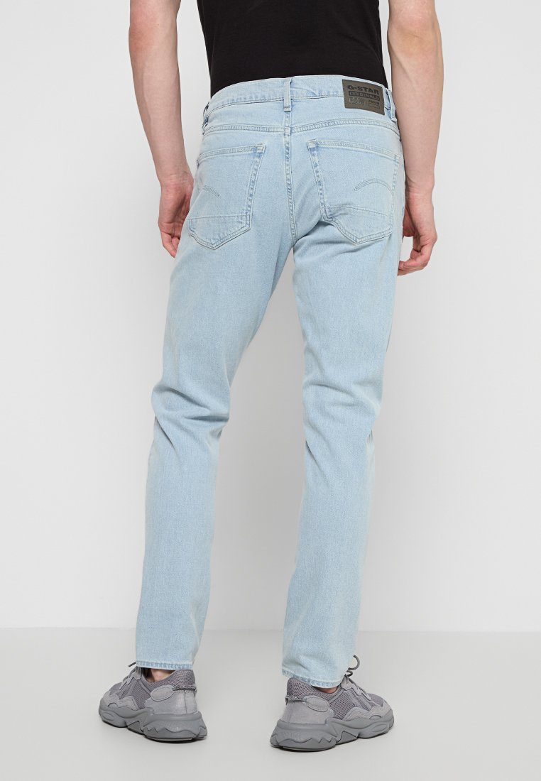 Helleblaue Jeans mit geradem Bein, mit Gesäßtaschen und einem Patch am Bund; getragen mit grauen Turnschuhen.