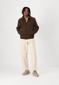 Veste zippée marron avec un col, ourlet élastique et poches latérales, associée à un pantalon beige clair et des chaussures beiges.