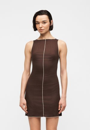 Žena s krátkymi tmavými vlasmi, oblečená v priliehavých hnedých šatách bez rukávov s bielymi vertikálnymi a horizontálnymi stehovými detailmi.