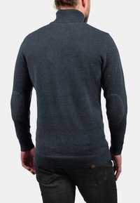 Dunkelblauer Pullover mit hohem Kragen, mit gerippten Ärmelbündchen und Saum. Auffällige Ellbogenpatches und weiche Textur. Rückansicht gezeigt.