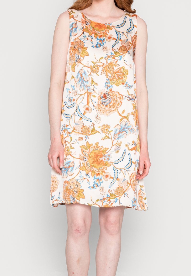 Robe sans manches longueur genoux avec des motifs floraux orange, bleu et beige sur fond blanc portée par une personne debout.