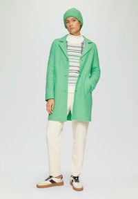 Manteau en laine vert avec un design à col, fermeture par boutons et poches latérales, porté sur un col roulé rayé et un pantalon crème. Baskets noir et blanc.