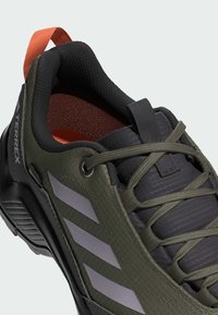 Zapato de trail Terrex con un upper texturizado de color verde oliva, acentos en negro, rayas reflectantes y una pestaña de tirón naranja en el talón.